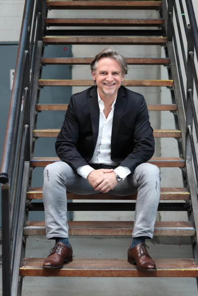 Peter Ehlers, Aufsichtsrat der Luana AG sitzt gekleidet in Jeans und Sakko auf einer Treppe, lächelt freundlich in die Kamera und hat dabei die Hände gefaltet.